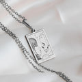 Silver pendant necklace featuring a design of two facing skulls on a rectangular card shape with a detailed line drawing. The necklace is shown against a white background, highlighting the engraved details.
