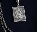 Silver-toned pendant with an engraved design of two regal women holding a heart. The item is suspended from a chain and positioned against a dark blue background.