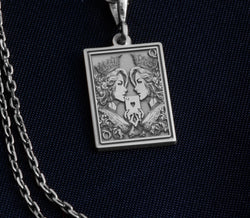 Silver-toned pendant with an engraved design of two regal women holding a heart. The item is suspended from a chain and positioned against a dark blue background.