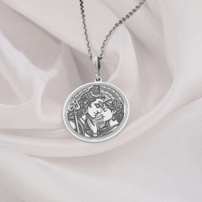 Silver toned circular pendant showing a detailed relief carving of Shiva and Parvati. This necklace features a silver chain and is displayed against a dark blue fabric background.