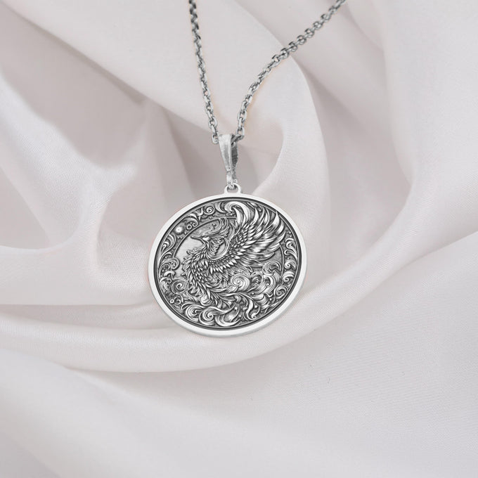 Silver-toned circular pendant featuring a detailed eagle design encircled by decorative scrollwork. The pendant is suspended from a silver chain and rests on soft white material.