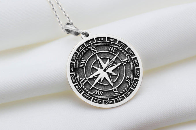 Silver compass directional pendant with a circular dial design featuring cardinal directions and a central cross motif. The pendant has a polished surface finish and is shown from the front view, attached to a silver chain.