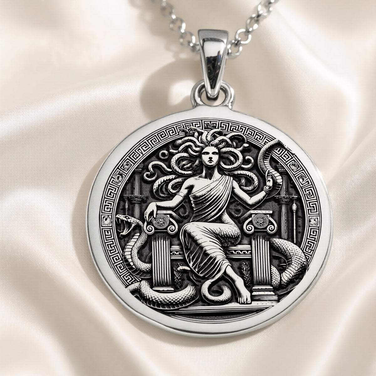 Silver circular pendant featuring an intricate design of a mythological figure entwined with a serpentine creature. The pendant is attached to a silver chain and rests on a dark blue fabric background.