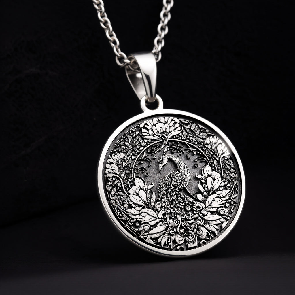 Silver colored circular pendant featuring an intricate embossed design of a peacock surrounded by detailed foliage. The pendant is attached to a silver-toned chain and displayed on a textured surface. - View 11