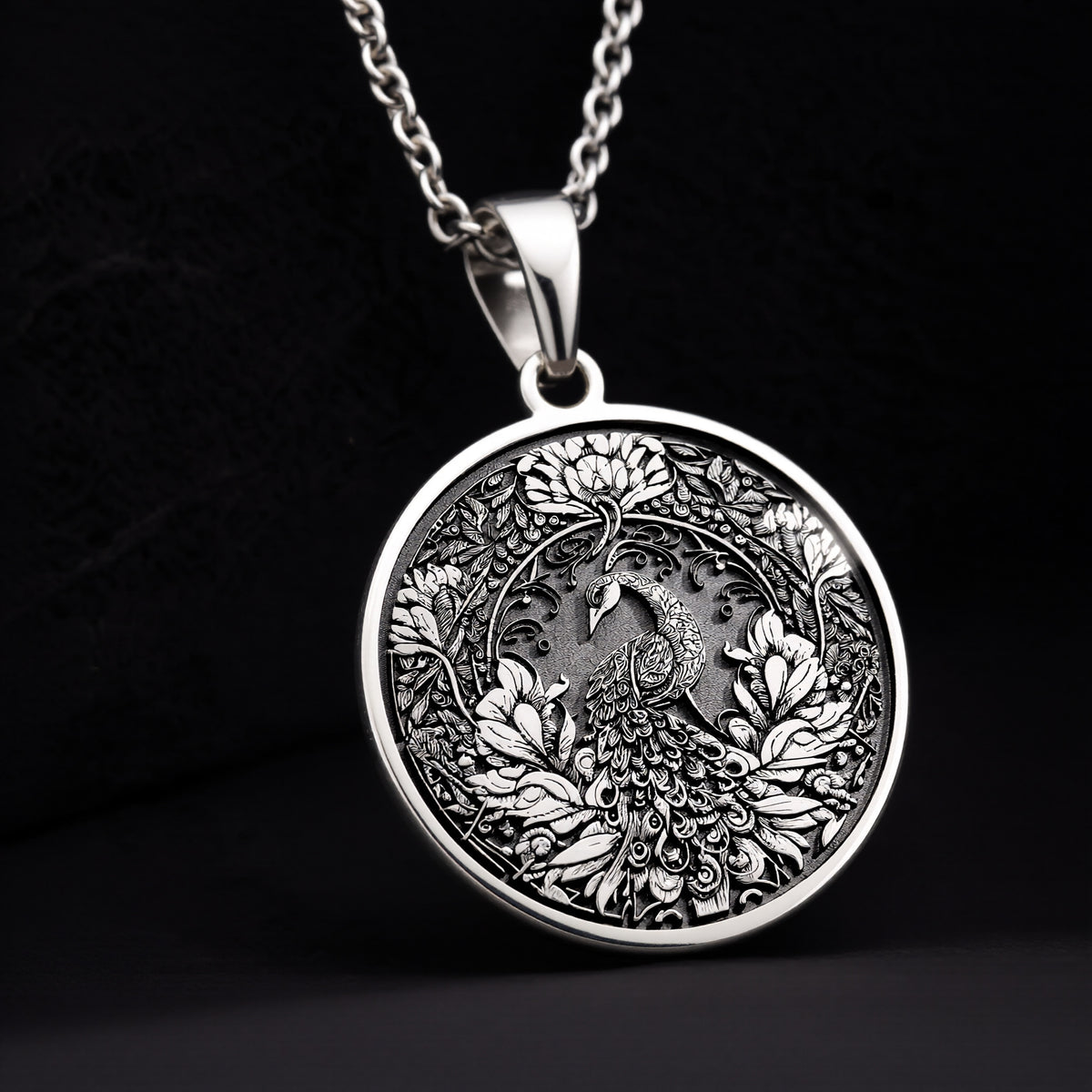 Silver colored circular pendant featuring an intricate embossed design of a peacock surrounded by detailed foliage. The pendant is attached to a silver-toned chain and displayed on a textured surface. - View 11
