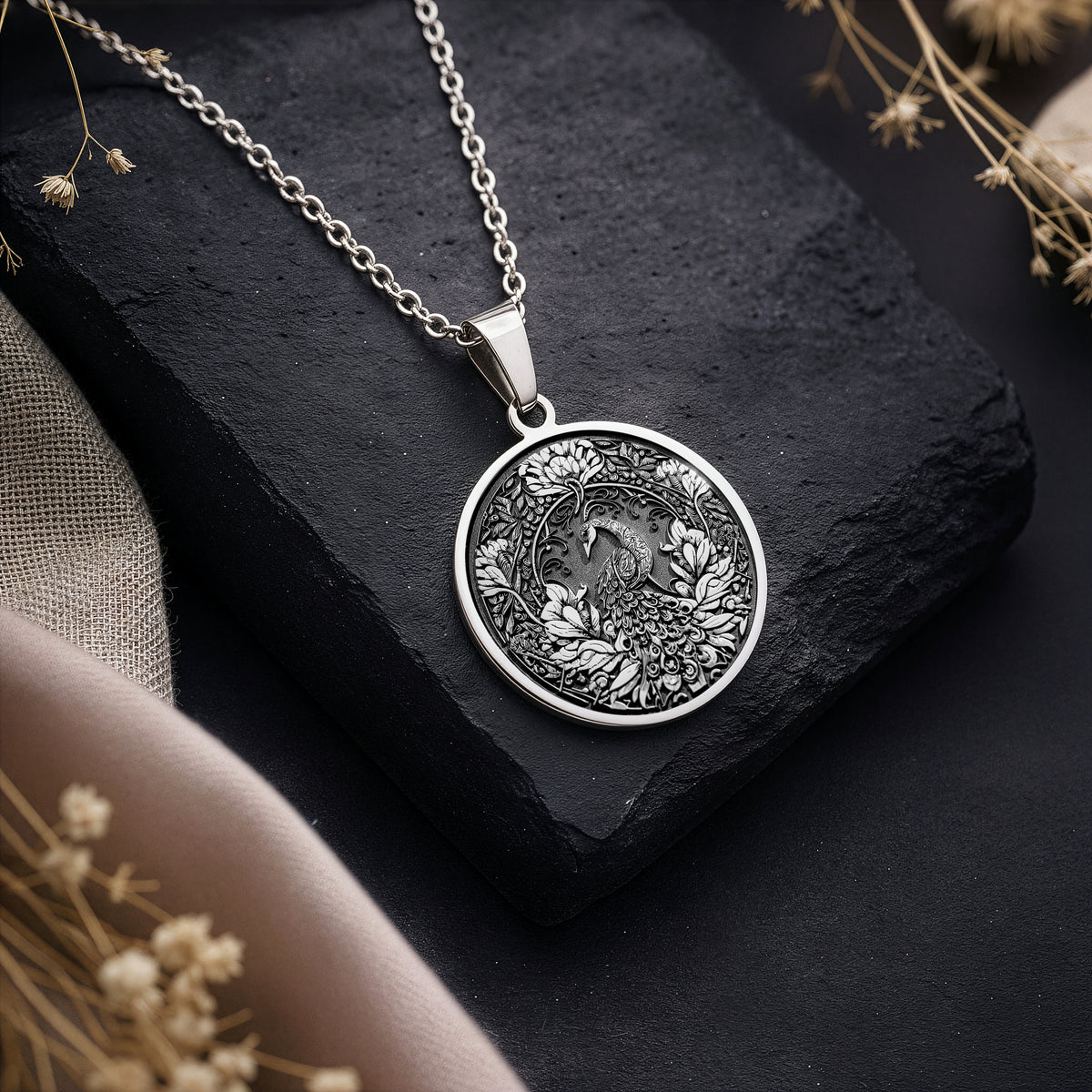Silver colored circular pendant featuring an intricate embossed design of a peacock surrounded by detailed foliage. The pendant is attached to a silver-toned chain and displayed on a textured surface. - View 3