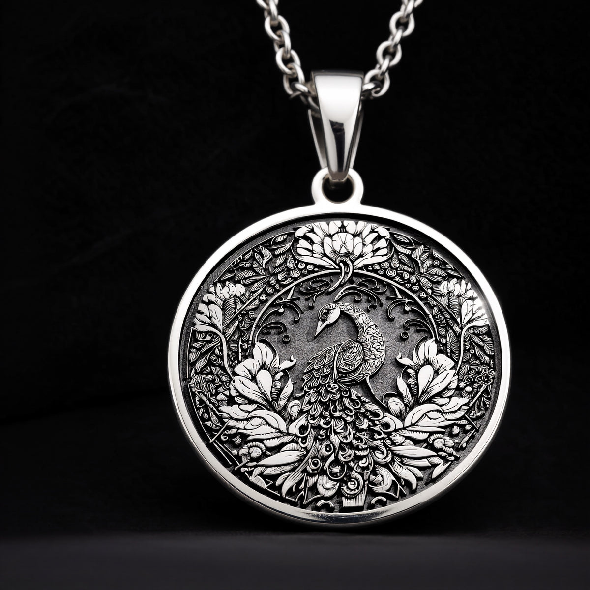 Silver colored circular pendant featuring an intricate embossed design of a peacock surrounded by detailed foliage. The pendant is attached to a silver-toned chain and displayed on a textured surface. - View 8