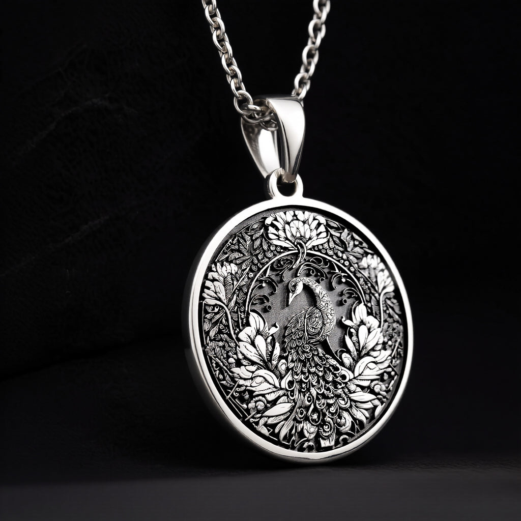 Silver colored circular pendant featuring an intricate embossed design of a peacock surrounded by detailed foliage. The pendant is attached to a silver-toned chain and displayed on a textured surface. - View 9