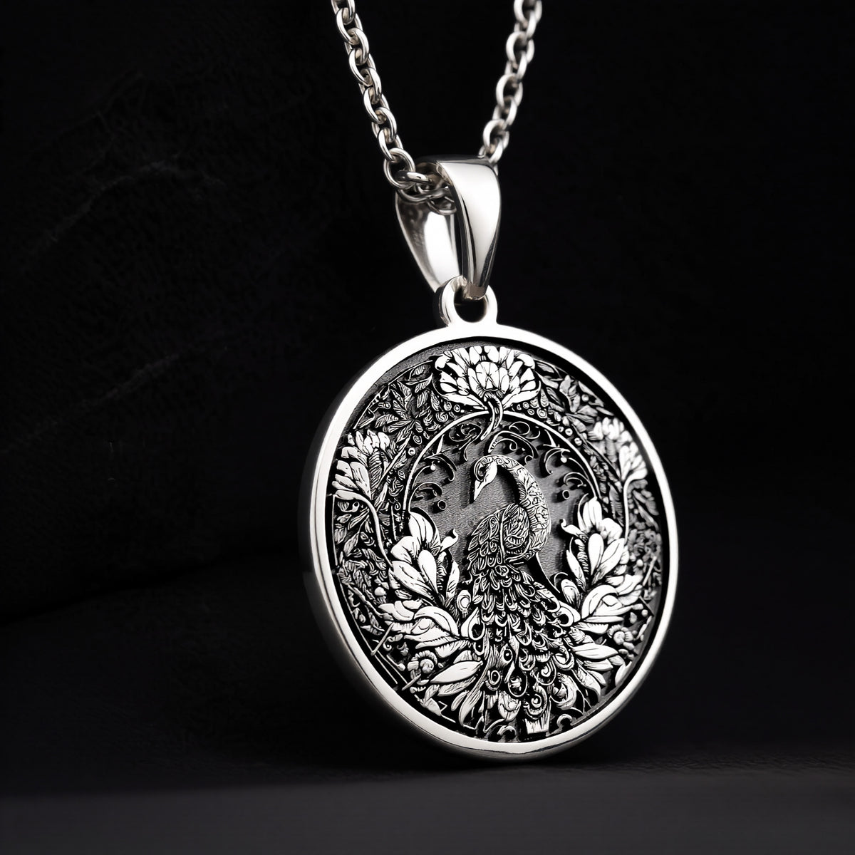 Silver colored circular pendant featuring an intricate embossed design of a peacock surrounded by detailed foliage. The pendant is attached to a silver-toned chain and displayed on a textured surface. - View 9