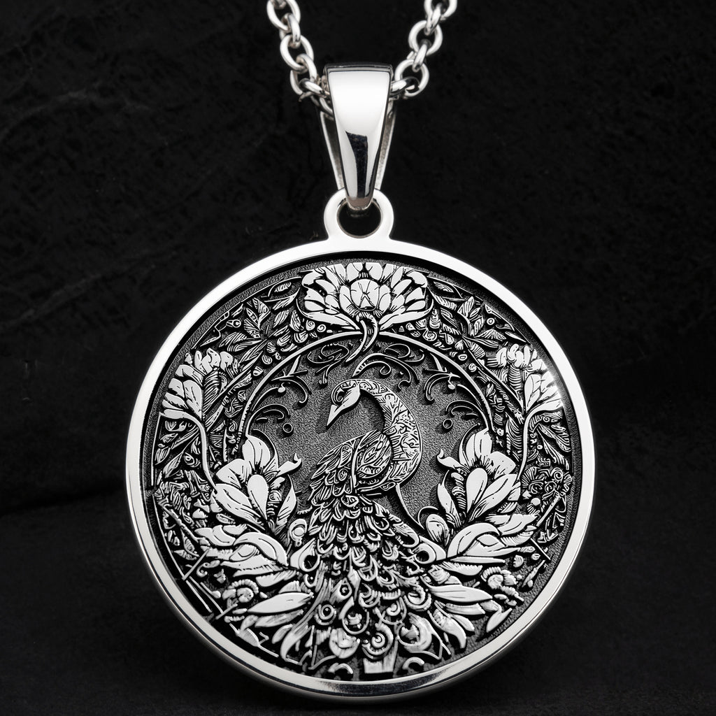 Silver colored circular pendant featuring an intricate embossed design of a peacock surrounded by detailed foliage. The pendant is attached to a silver-toned chain and displayed on a textured surface.