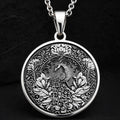 Silver colored circular pendant featuring an intricate embossed design of a peacock surrounded by detailed foliage. The pendant is attached to a silver-toned chain and displayed on a textured surface.