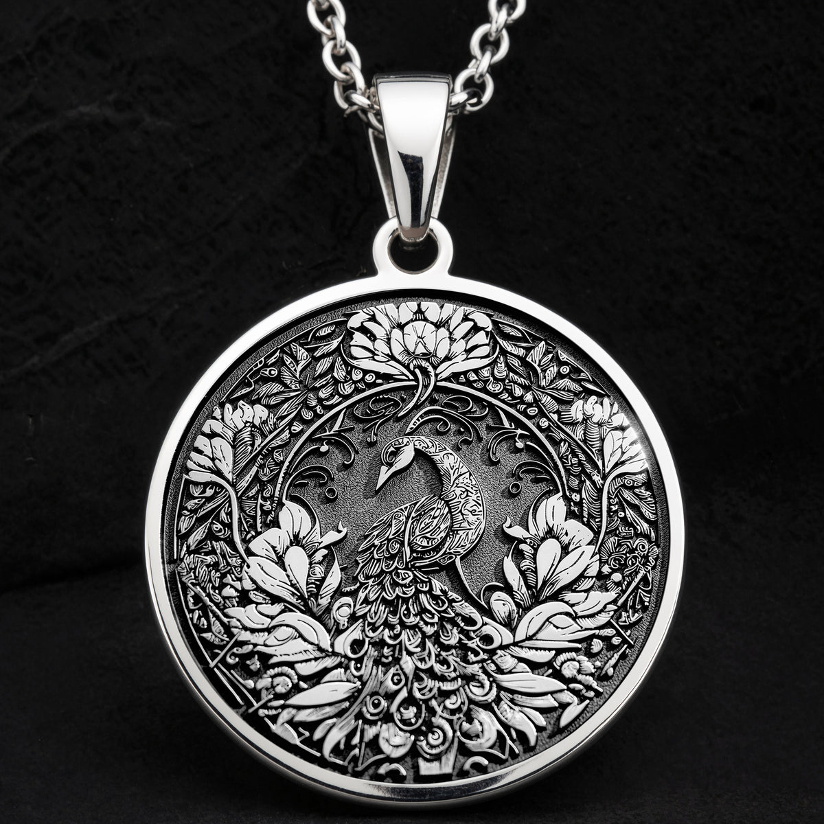 Silver colored circular pendant featuring an intricate embossed design of a peacock surrounded by detailed foliage. The pendant is attached to a silver-toned chain and displayed on a textured surface.