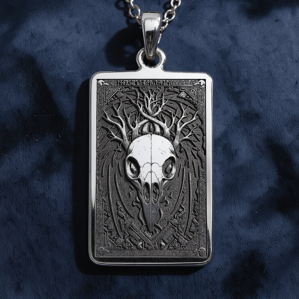 Silver pendant necklace displaying a detailed skull motif enclosed in an ornate frame. The necklace is laid out on black fabric with loose, colorful gemstone accents.