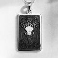 Silver pendant necklace featuring a detailed skull design with intricate framing. This silver-toned item is presented on a dark background alongside scattered faceted gemstones.