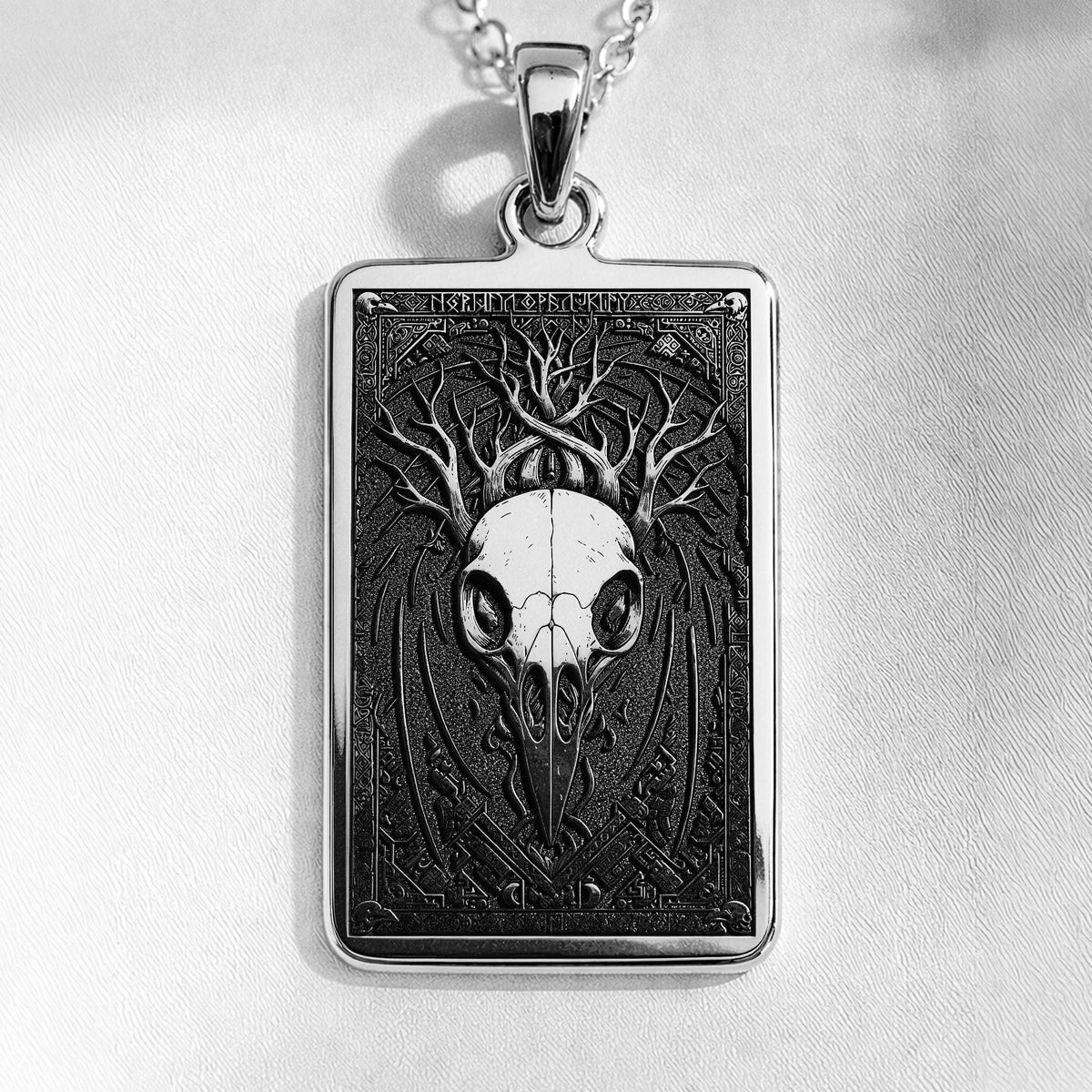 Silver pendant necklace featuring a detailed skull design with intricate framing. This silver-toned item is presented on a dark background alongside scattered faceted gemstones.
