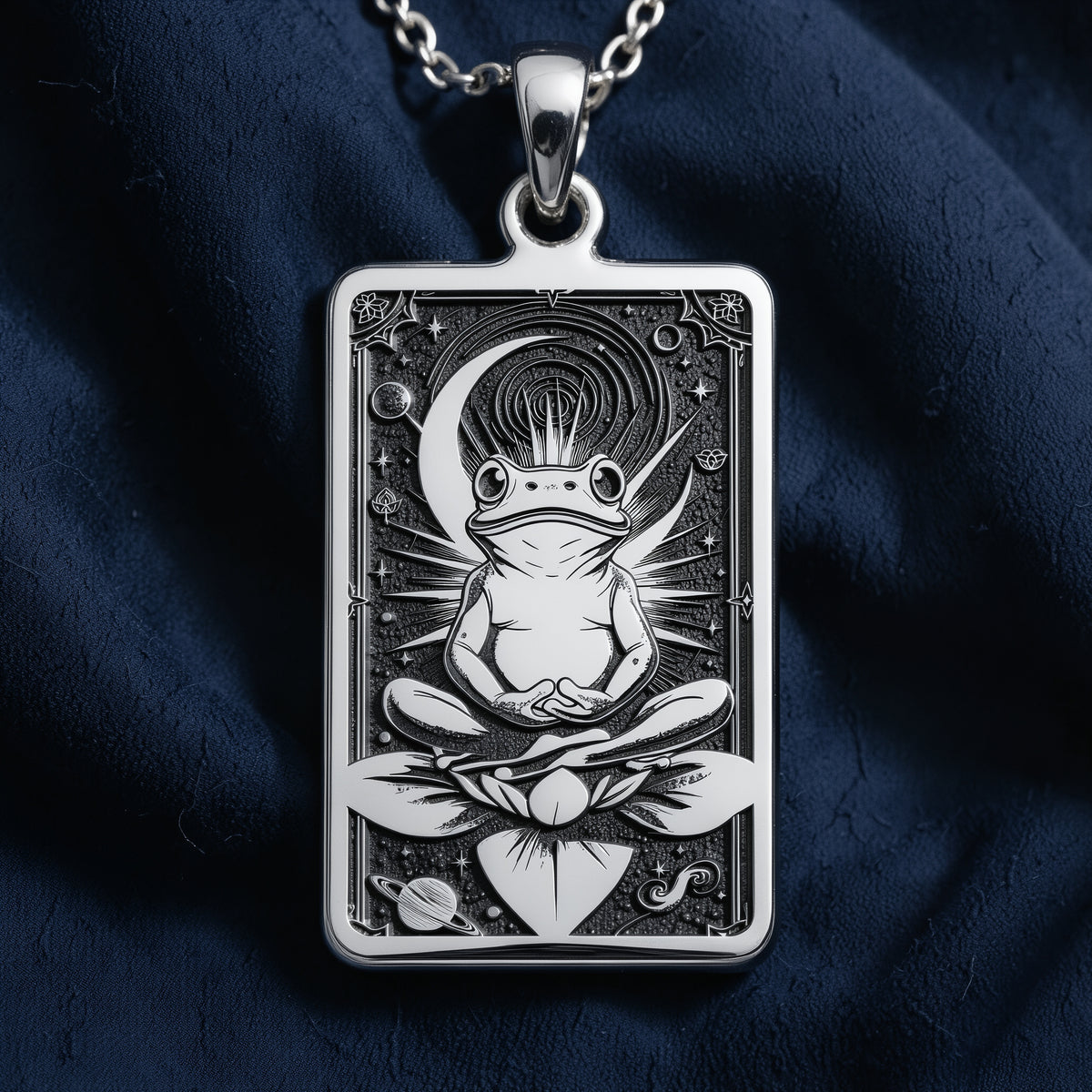 Silver rectangular pendant necklace displaying an engraved image of a frog resting among plants. The pendant has a smooth, polished surface and is arranged with various cut gemstones on a dark fabric setting.
